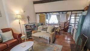 En hyggelig stue med en sofa, et sofabord og en pejs. Der er et bogskab med bøger og en lampe. Værelset har trægulv og en trappe, der fører til en etage ovenover.