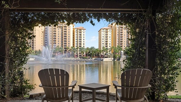 A covered outdoor seating area with two Adirondack chairs and a small table, overlooking a body of water with a fountain and high-rise buildings in the background. - 3BD/Orlando Near Disney - 5 Pools, Game Room, Café and Restaurant On-Site! (Lake Buena Vista)