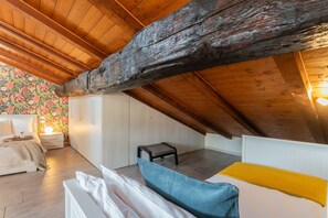 Un dormitorio acogedor en el ático, con techo inclinado, vigas de madera, una cama con manta amarilla, una mesita de noche con lámpara y un taburete pequeño.