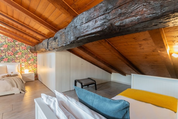 A cozy attic bedroom with a sloped ceiling, wooden beams, a bed with a yellow blanket, a nightstand with a lamp, and a small stool.