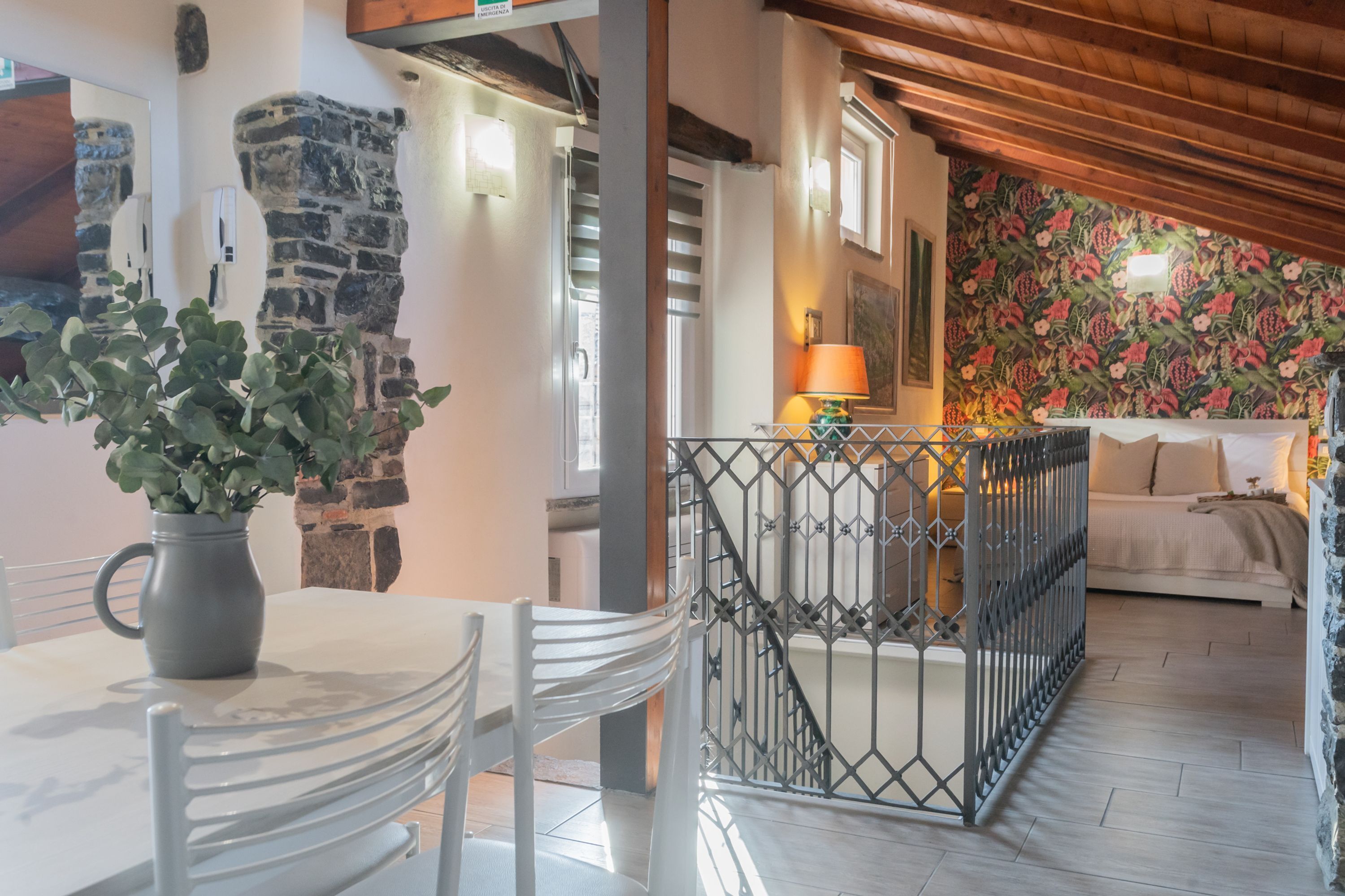 Un comedor con una mesa y sillas, una planta en maceta, un muro de piedra y una escalera que lleva a una sala de estar con un diseño floral en la pared.
