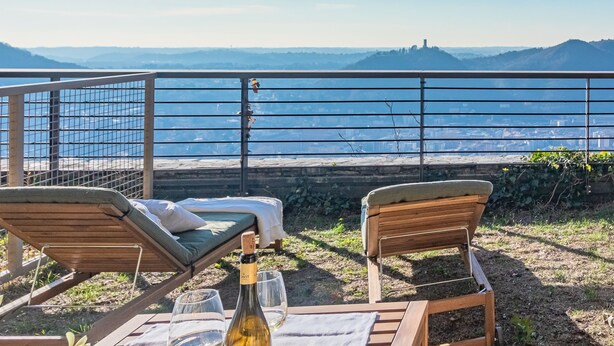 A terrace with two wooden lounge chairs, a bottle of wine, and glasses on a table, overlooking a scenic view of the sea and distant hills. - Aurora Apartment in Como (Como)