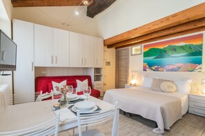 Une chambre d’hôtel moderne avec un lit, une table à manger entourée de chaises blanches, un canapé rouge et un tableau représentant un paysage accroché au mur.