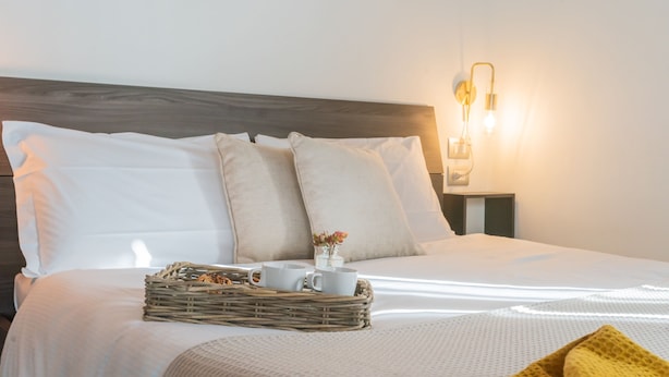 A neatly made bed with white linens, a wicker tray with two white mugs, and a small vase with flowers on the bed.