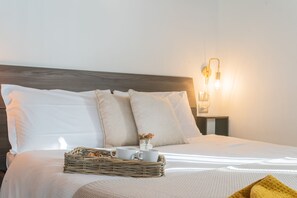 Een netjes opgemaakt bed met witte lakens, een rieten dienblad met twee witte mokken en een kleine vaas met bloemen op het bed.