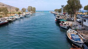 Una marina con varias embarcaciones amarradas a lo largo de la orilla, un muelle con vehículos, y un fondo de árboles y colinas.