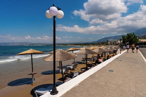 Une promenade en bord de mer, agrémentée de chaises longues et de parasols en chaume, un sentier pavé et une vue sur la mer et les montagnes lointaines.