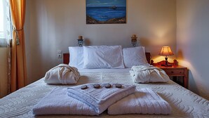 Habitación de hotel con cama, ropa de cama blanca, un cuadro de una escena marina, una mesita de noche con lámpara y una ventana con cortinas.