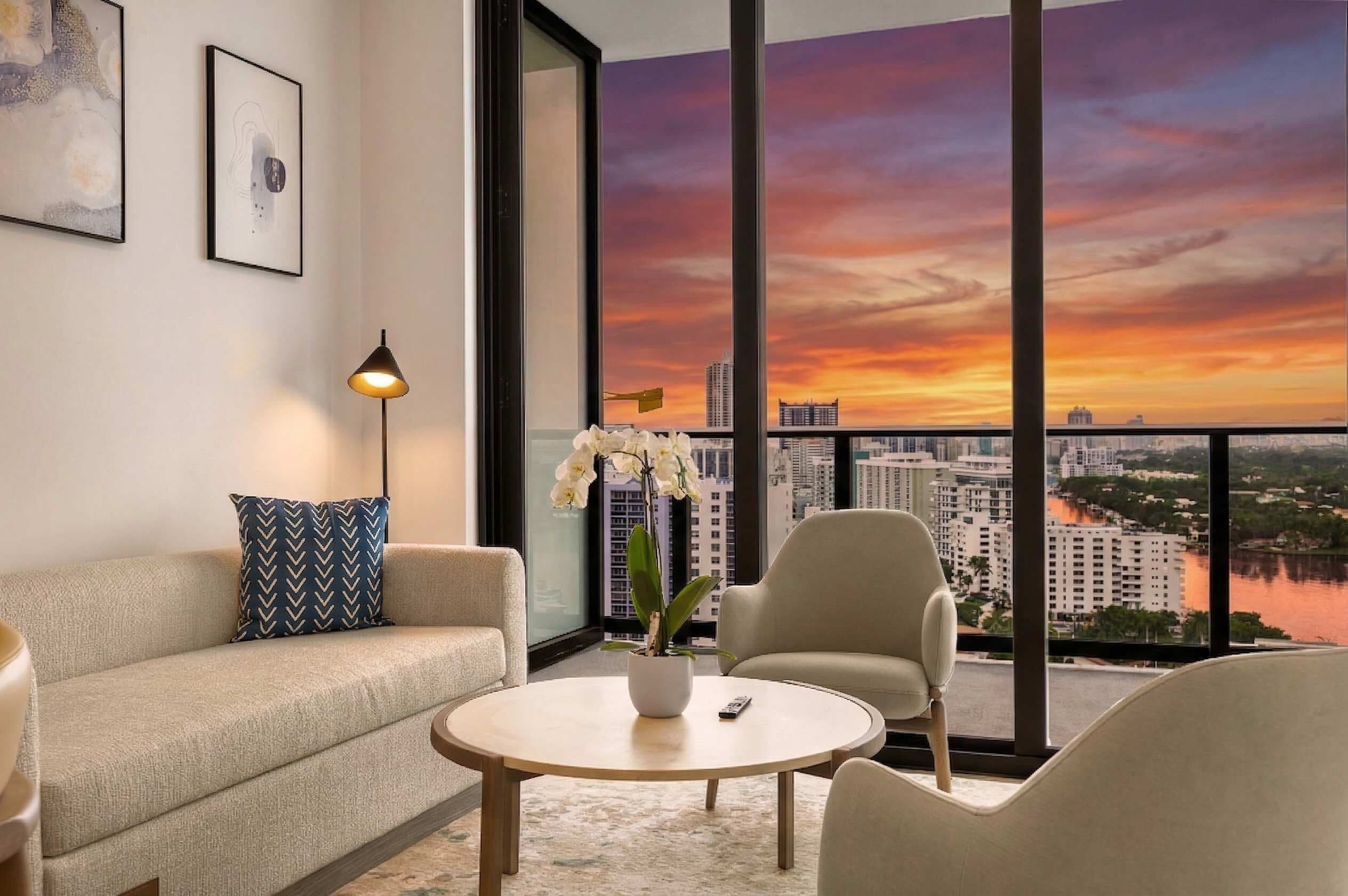 Uma sala de estar moderna com sofá, poltrona e mesa de centro, janelas amplas com vista para a cidade e uma orquídea em vaso.