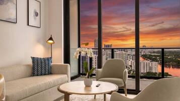 Uma sala de estar moderna com sofá, poltrona e mesa de centro, janelas amplas com vista para a cidade e uma orquídea em vaso.