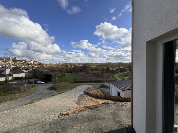 A view from a window overlooking a gravel driveway, a white building, and a distant town with a church tower.