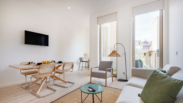 A modern living room with a sofa, a dining table with chairs, a TV mounted on the wall, and a large window offering a view of the outside. - The Superior Green at BoaVista Flats (Porto)