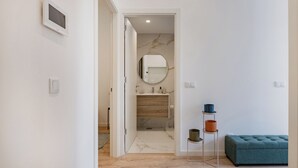 A modern bathroom with a marble wall, a round mirror, a wooden vanity, and a blue ottoman. - The Superior Green at BoaVista Flats (Porto)