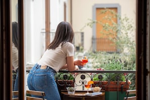 Eine Frau sitzt an einem Tisch im Freien mit einem Getränk, umgeben von Blumentöpfen und Blick auf ein Gebäude.