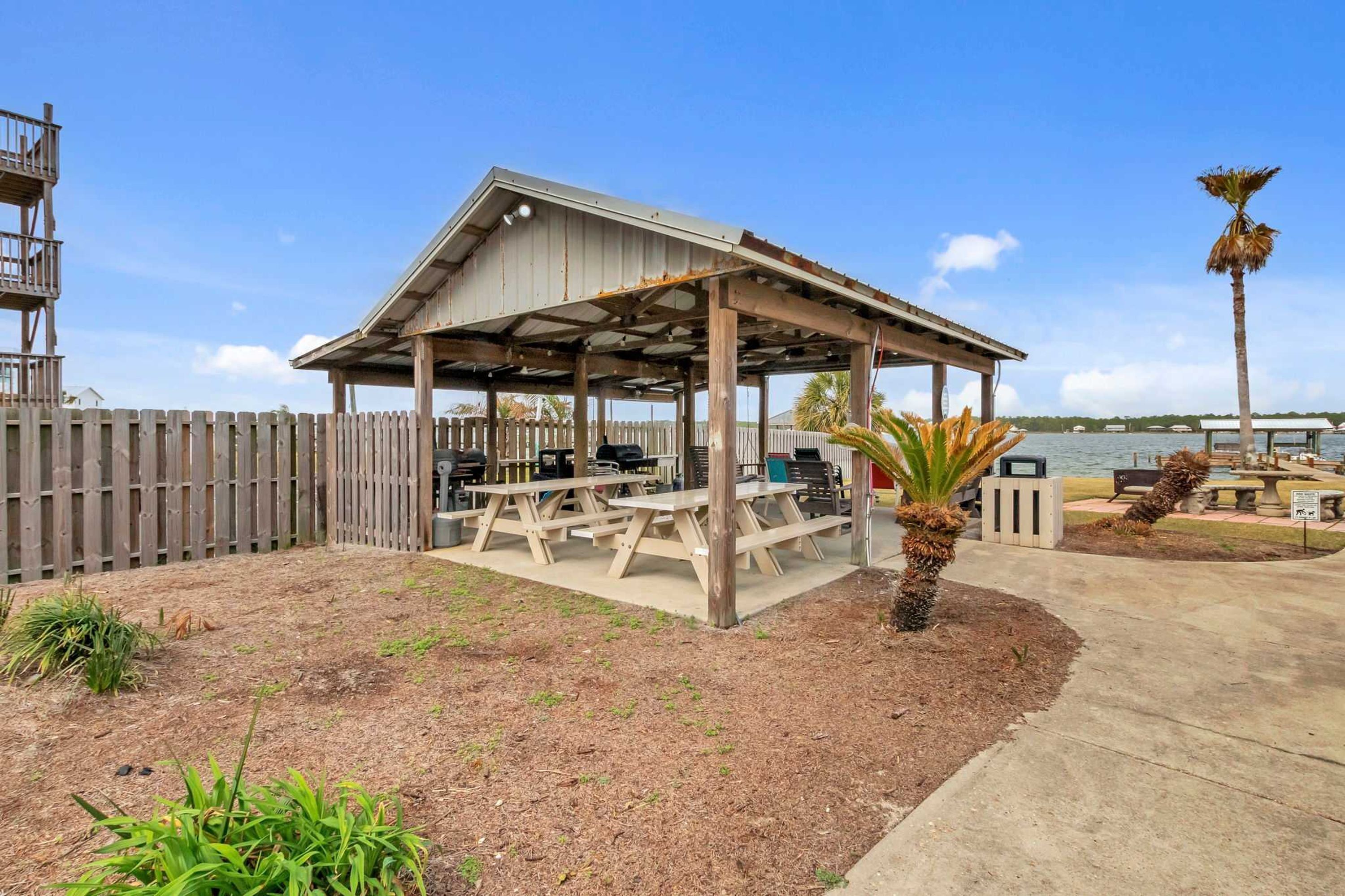 A covered outdoor seating area with picnic tables, a wooden fence, and a palm tree.