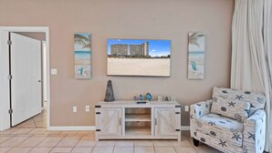 Un salon avec un mur de couleur beige, un téléviseur fixé au-dessus d’une console en bois, un canapé aux motifs évoquant la plage et une porte sur le côté gauche.