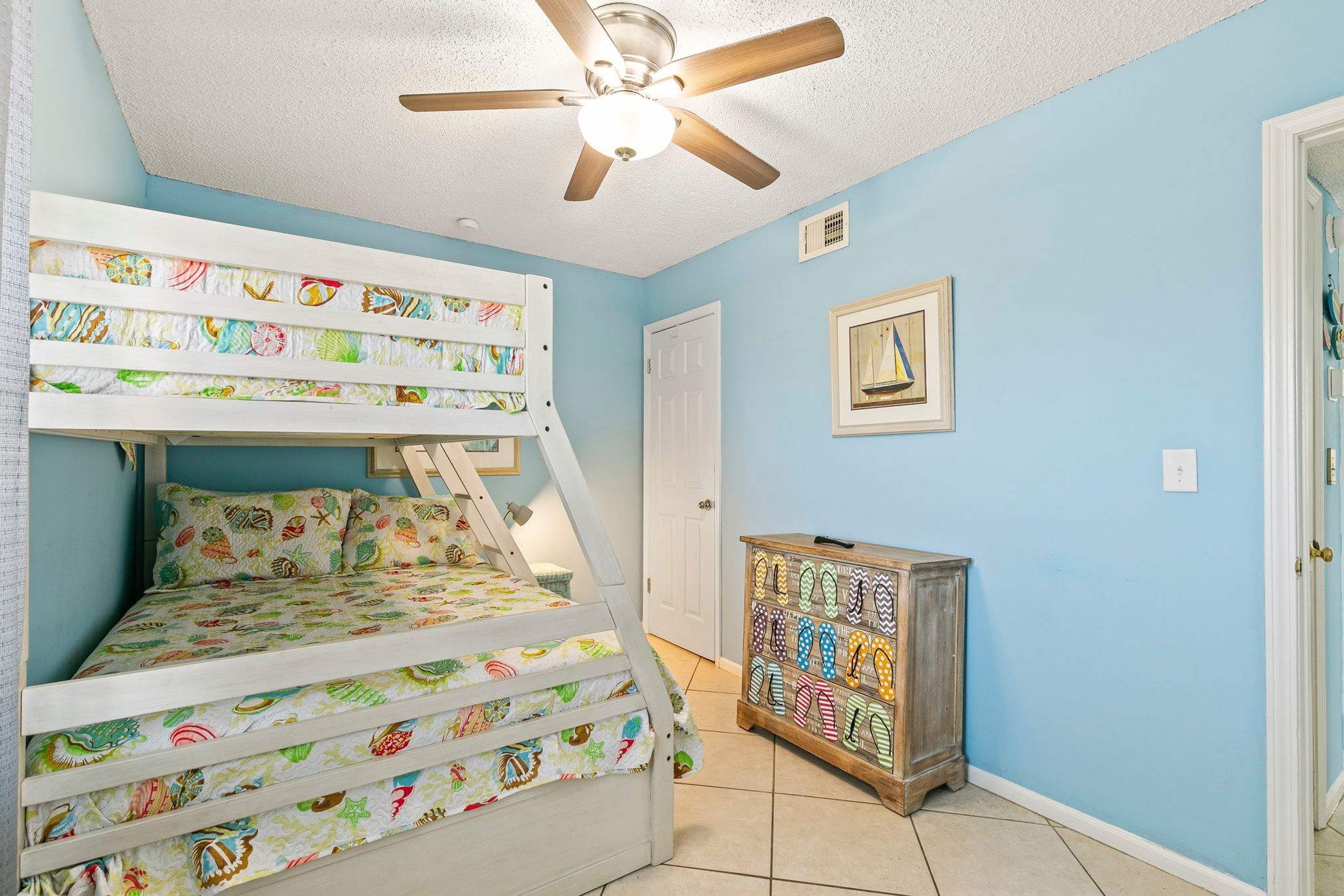 A room with a bunk bed, a ceiling fan, a door, a cabinet with decorative knobs, and a framed picture on the wall.