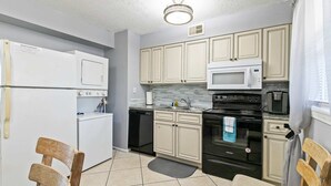 Una cocina compacta con armarios blancos, un microondas, un lavavajillas y una estufa. Hay dos sillas de madera y una alfombra pequeña en el suelo.