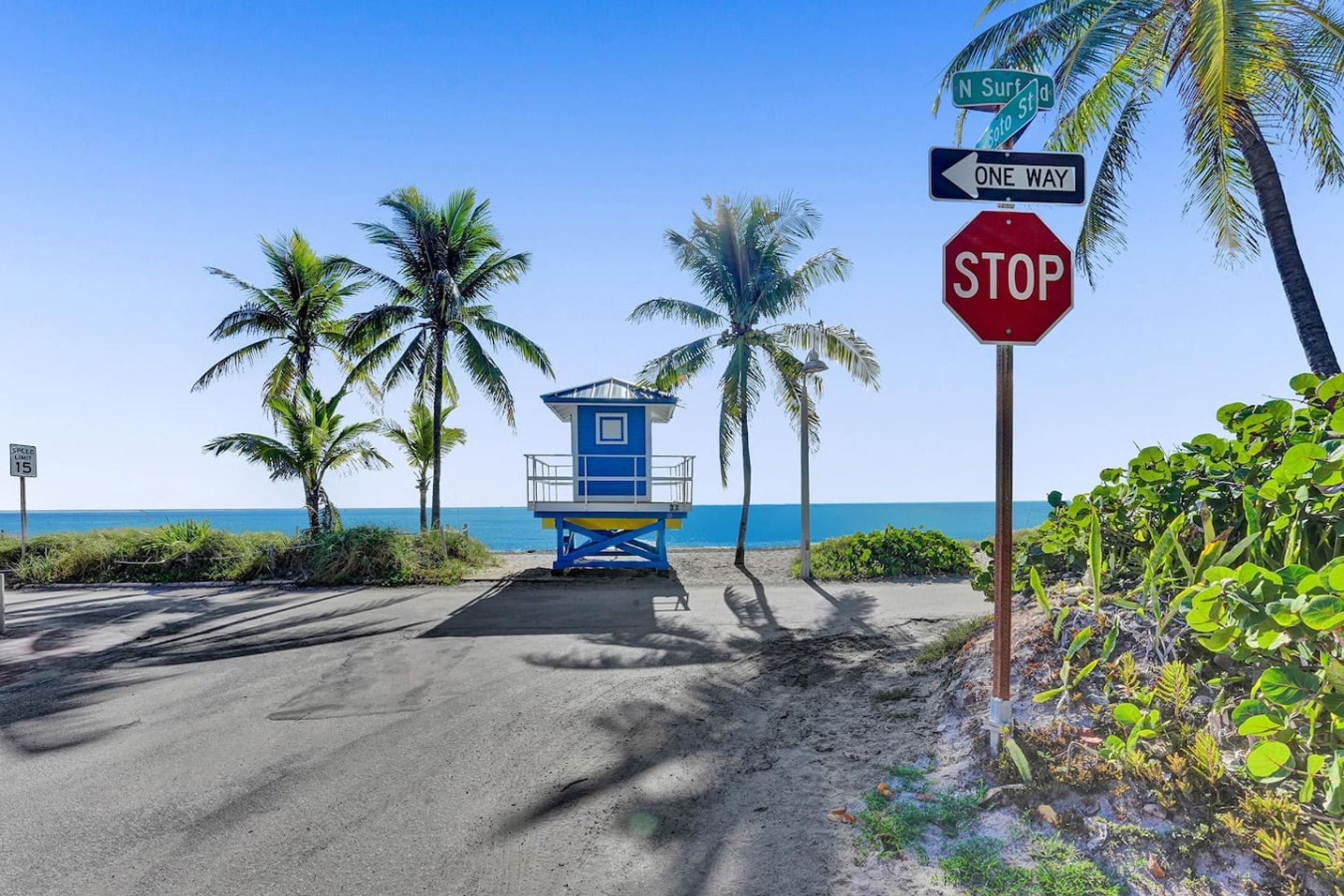 Una carretera frente a la playa, con una torre de salvavidas, palmeras y una señal de parada.