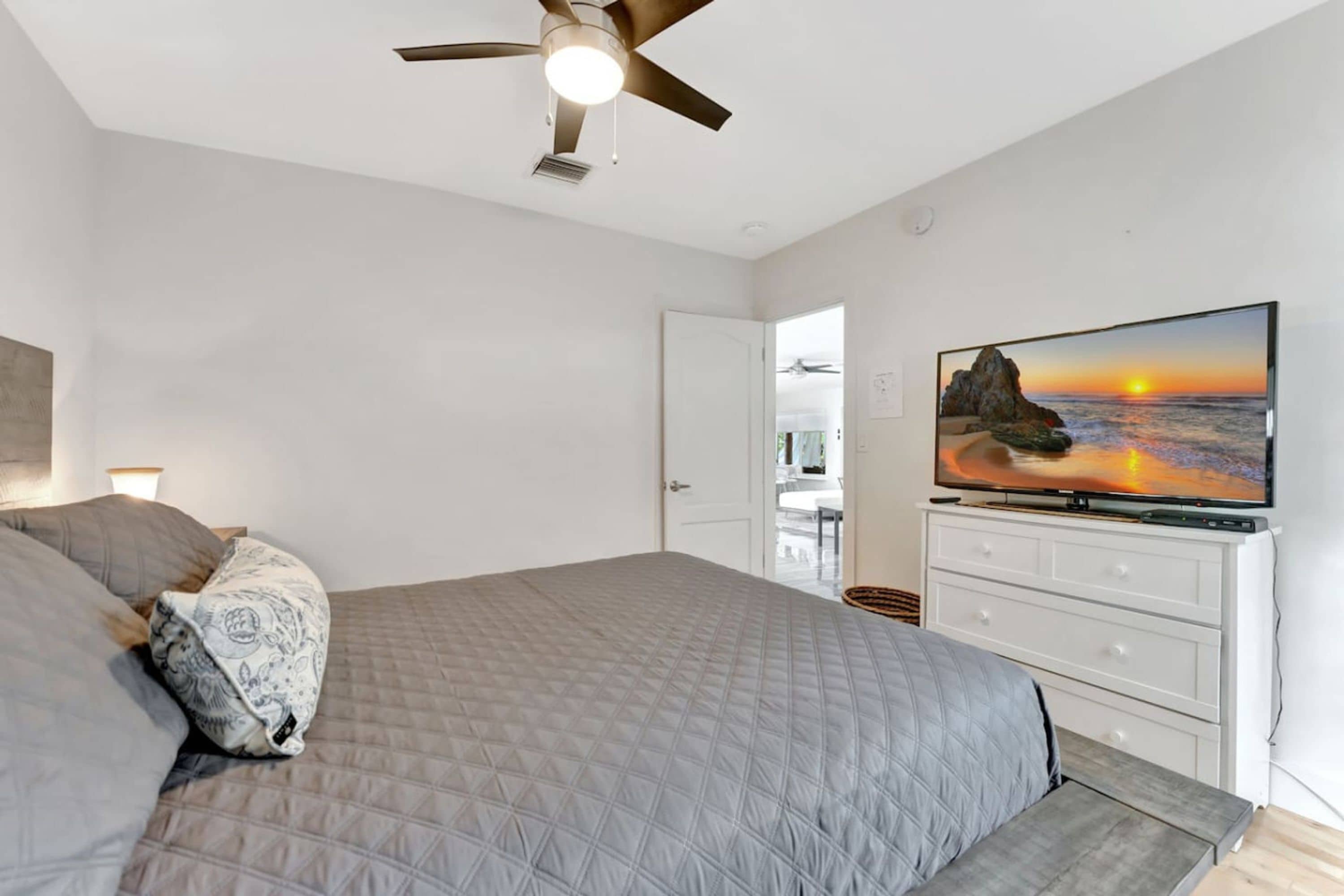A bedroom with a bed, a ceiling fan, a TV on a dresser, and a door leading to another room.