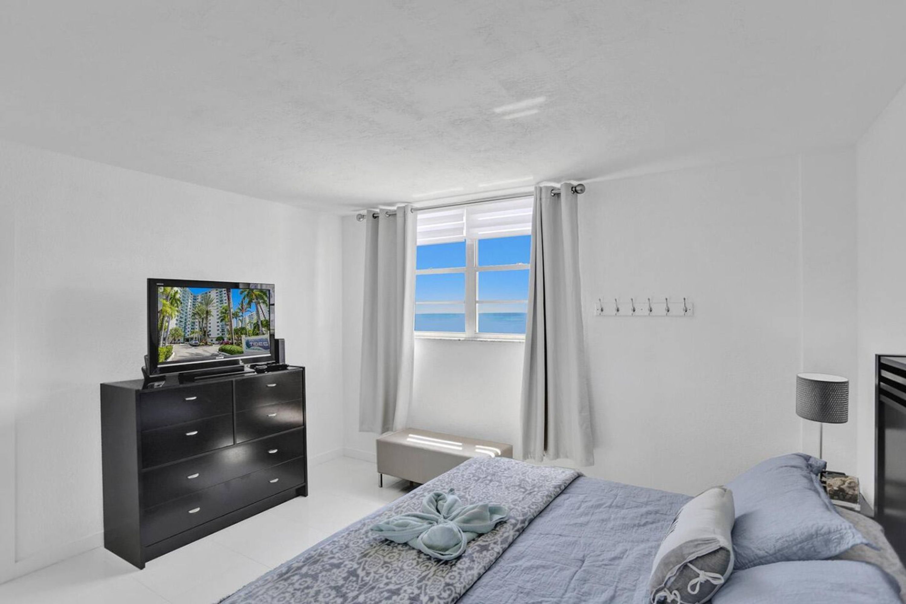 A modern bedroom with a bed, a TV on a black dresser, a window with curtains, and a bedside lamp.