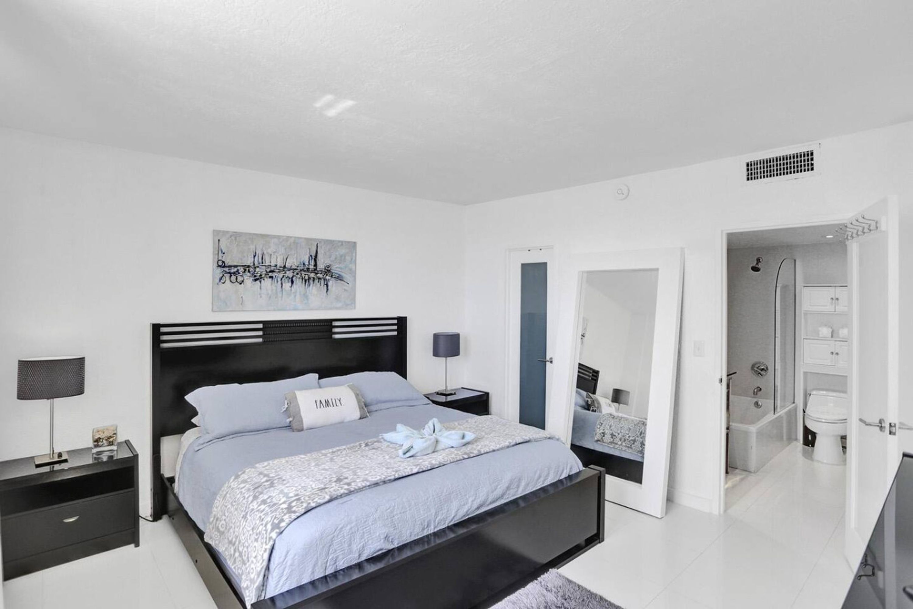 Une chambre moderne avec un lit, des tables de chevet, une commode, une œuvre d’art accrochée au mur et une salle de bain visible à travers une porte ouverte.