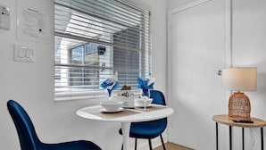 Un comedor con una mesa redonda, dos sillas azules, una mesita auxiliar con lámpara y una ventana con persianas.