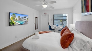 Una habitación de hotel con cama, ventilador de techo, televisor montado en la pared, ventana con persianas y un cuadro sobre la cama.
