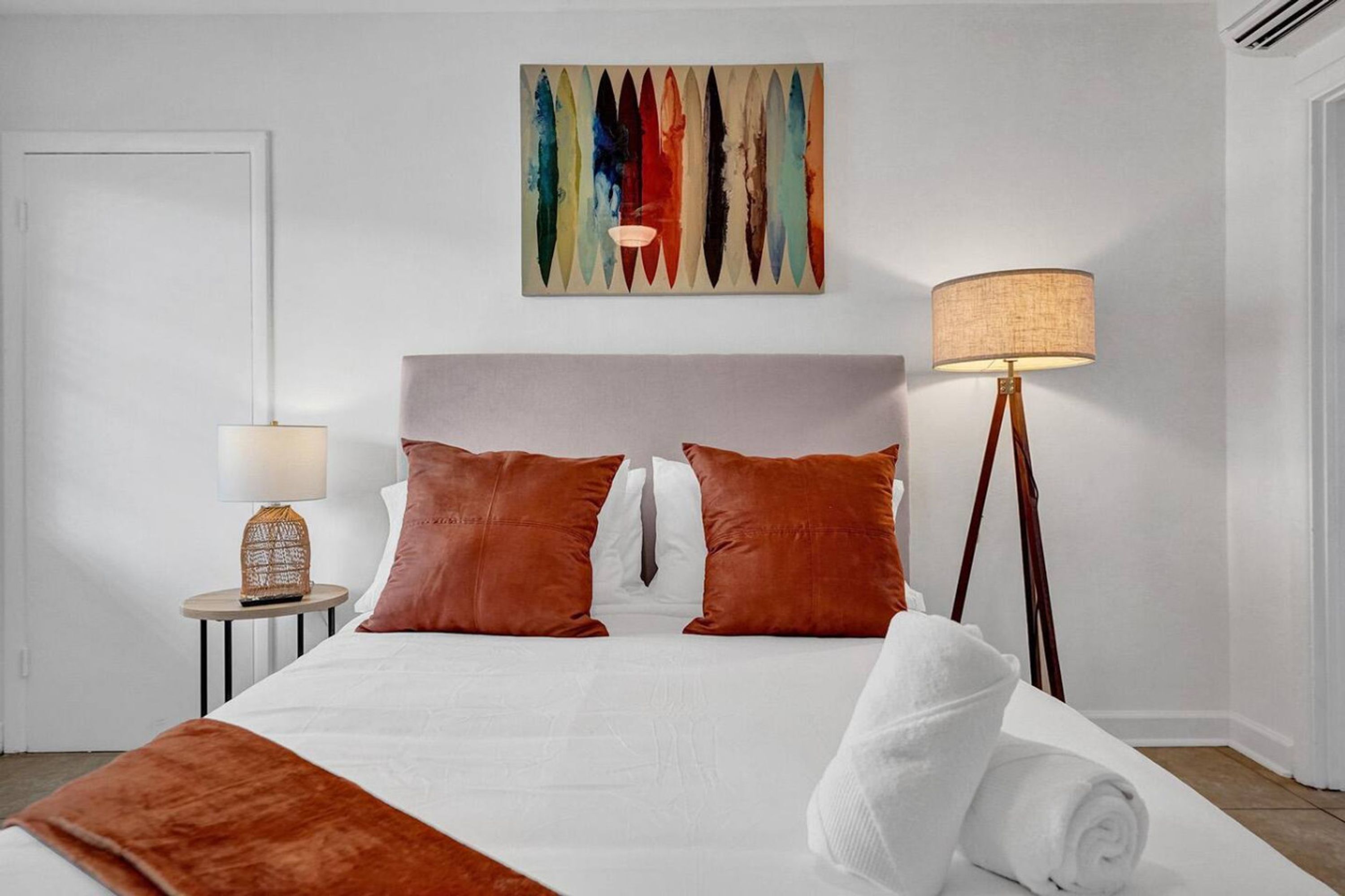 A neatly made bed with a white duvet and two brown pillows, a bedside lamp, and a colorful abstract painting on the wall.