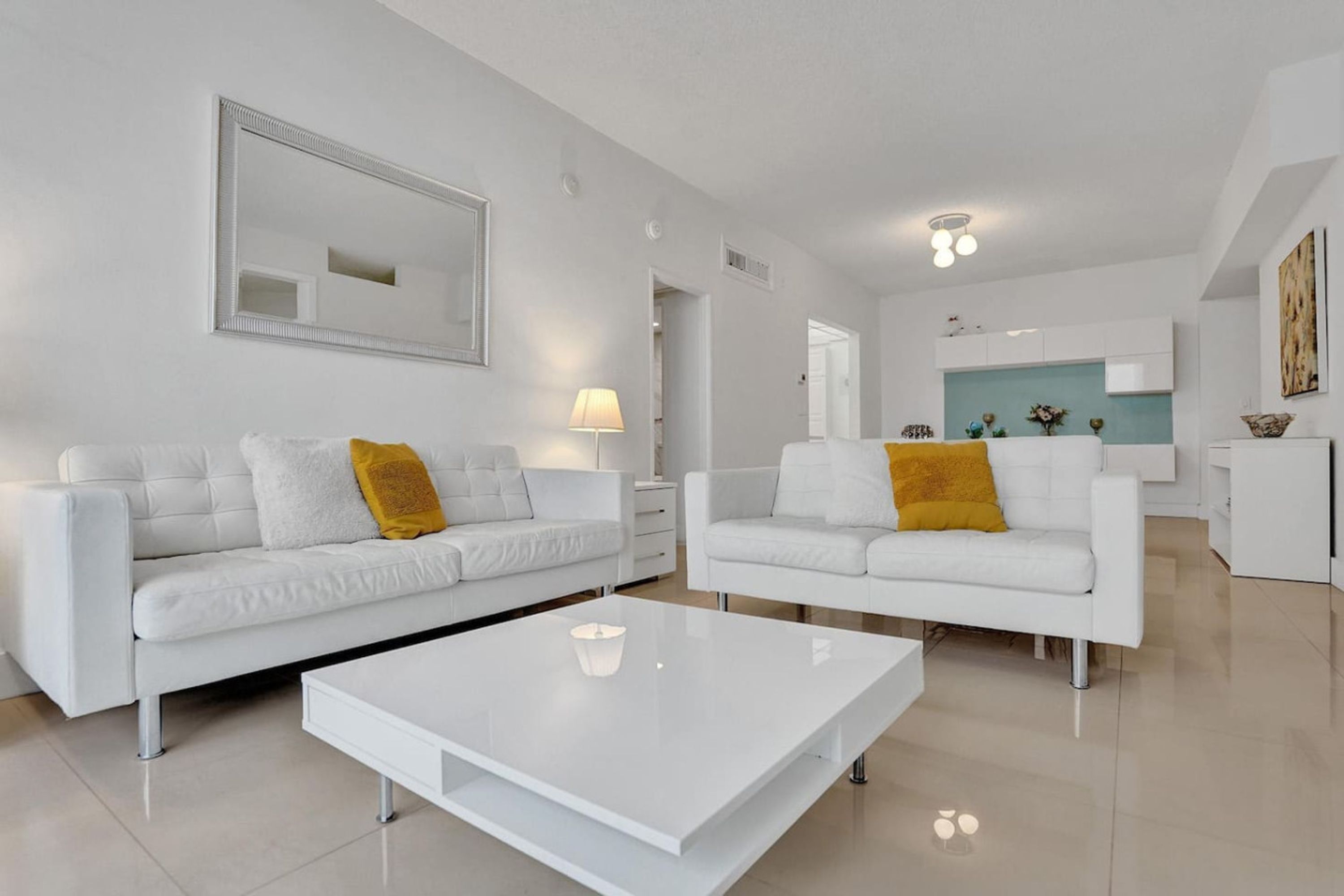 A modern living room with two white sofas, a white coffee table, and a kitchen in the background.