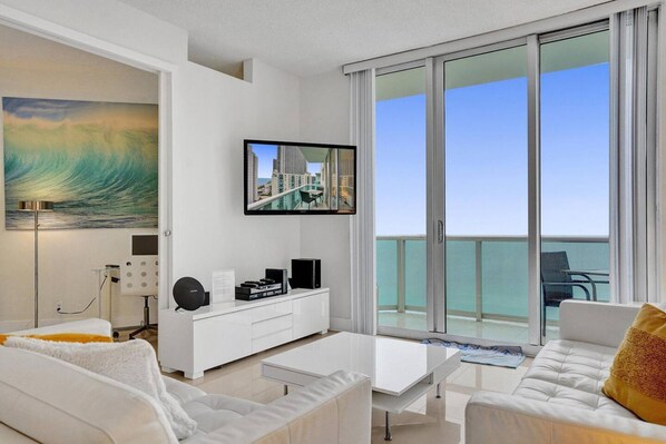 Una sala de estar moderna con un televisor grande, muebles blancos y un balcón con vista al océano.