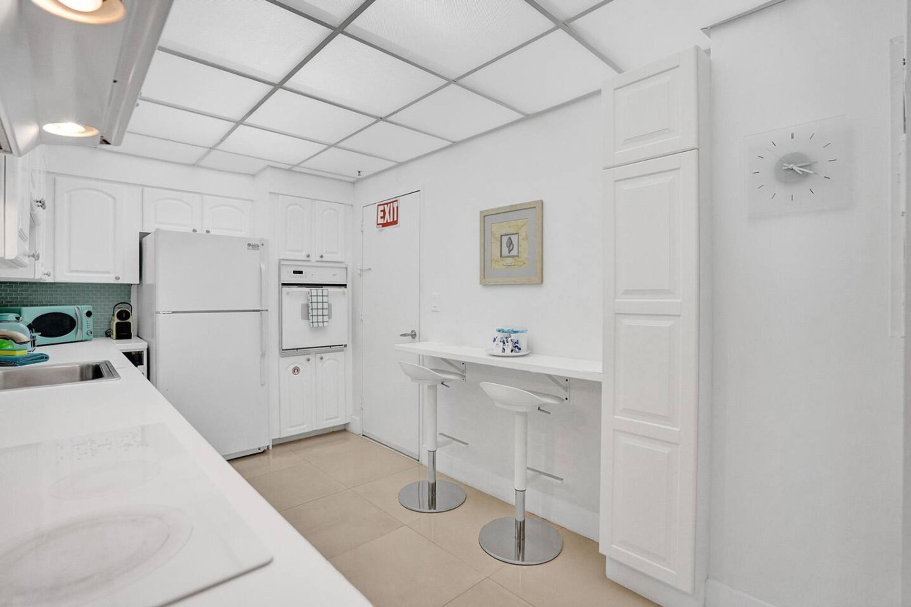 Cocina moderna con armarios blancos, horno empotrado, refrigerador y lavamanos. Hay dos taburetes de bar y un cuadro en la pared.