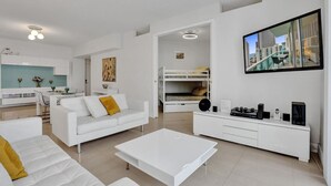 Ein modernes Wohnzimmer mit einem weißen Sofa, einem Couchtisch und einem an der Wand montierten Fernseher. Im Hintergrund ist eine Küchenzeile mit weißen Schränken und einem Esstisch zu sehen.