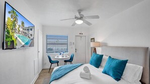 A hotel room with a bed, a ceiling fan, a TV displaying a pool view, a window with blinds, and a small table with chairs. - Waves on Desoto Beautiful Studio 8 (Hollywood)