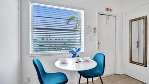 A small, bright room with a white table and two blue chairs, a window with blinds, an exit sign, and a door with a mirror. - Waves on Desoto Beautiful Studio 8 (Hollywood)