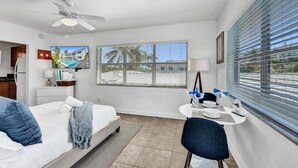 A hotel room with a bed, a desk with a chair, a ceiling fan, a window with blinds, and a view of palm trees and buildings. - Waves on Desoto Beautiful Studio (Hollywood)