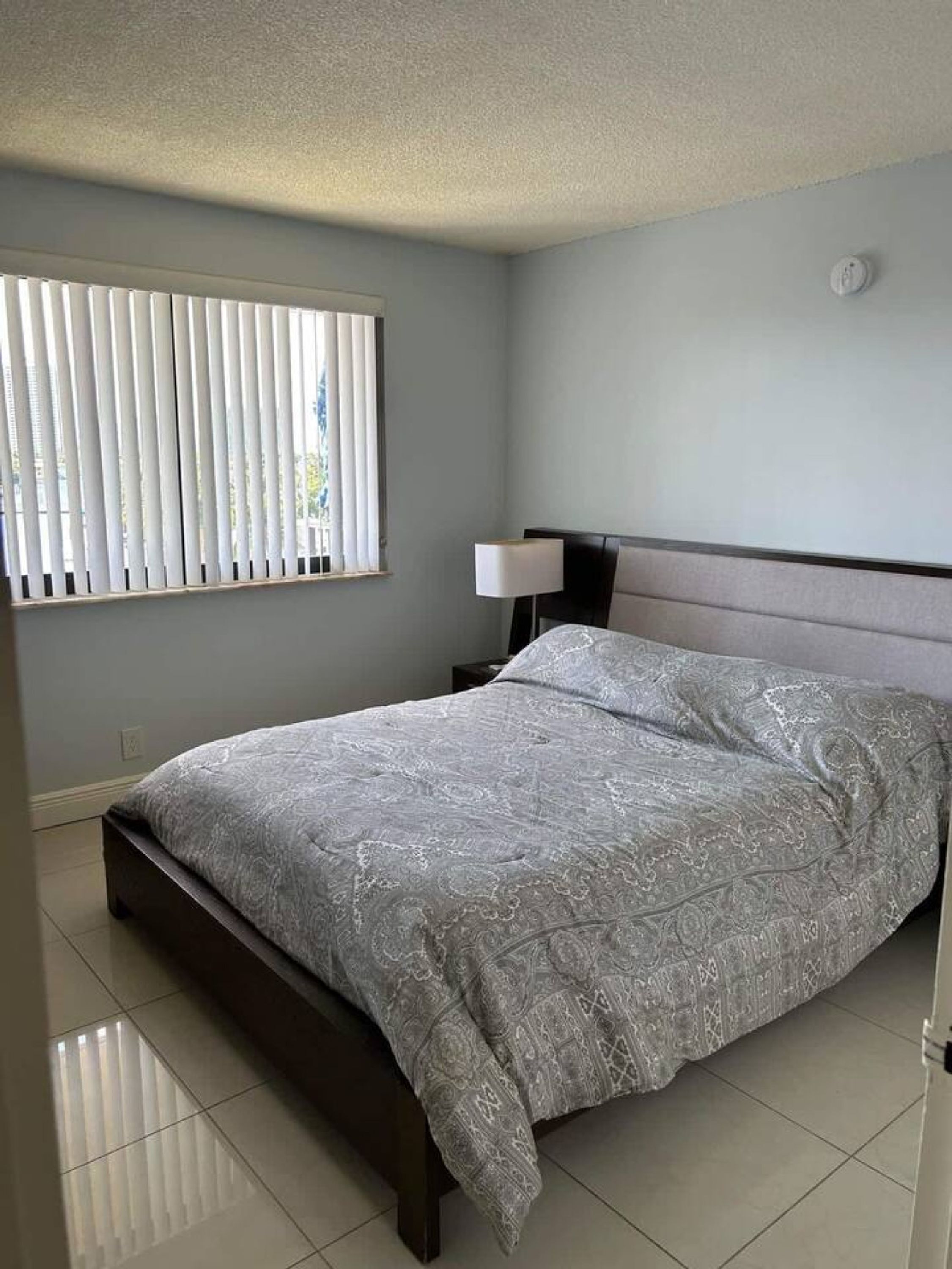 A bedroom with a bed, a nightstand, a lamp, and a window with blinds.