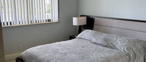 A bedroom with a bed, a nightstand, a lamp, and a window with blinds.