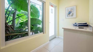 Habitación de hotel con un amplio ventanal corredera que da a un jardín tropical, un pequeño mostrador de recepción y una obra de arte enmarcada en la pared.