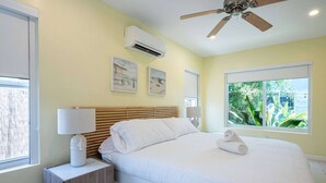 Un dormitorio con cama, mesitas de noche, ventilador de techo y ventana con vista a follaje.