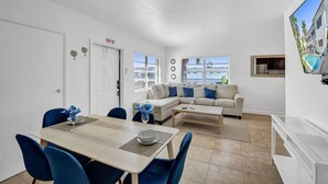 A modern living room with a dining area, a sofa set, a TV mounted on the wall, and a view of the outside.