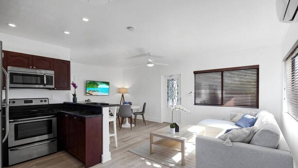 A modern kitchen with a microwave, stove, and dining area. There is a living room with a sofa and a TV. - Waves On Walnut 4-beach Access (Hollywood)