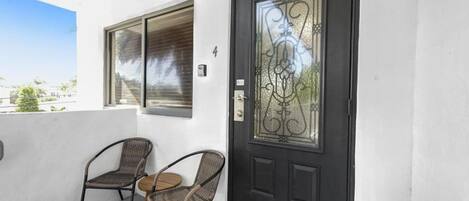 A black door with a decorative glass panel, two wicker chairs, and a small table on a balcony with a clear view of the sky and buildings in the background.