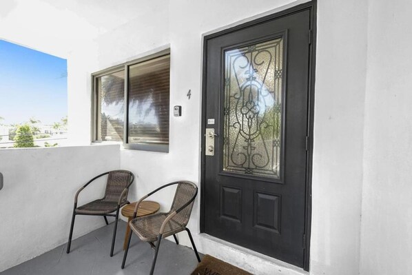 A black door with a decorative glass panel, two wicker chairs, and a small table on a balcony with a clear view of the sky and buildings in the background. - Waves On Walnut 4-beach Access (Hollywood)