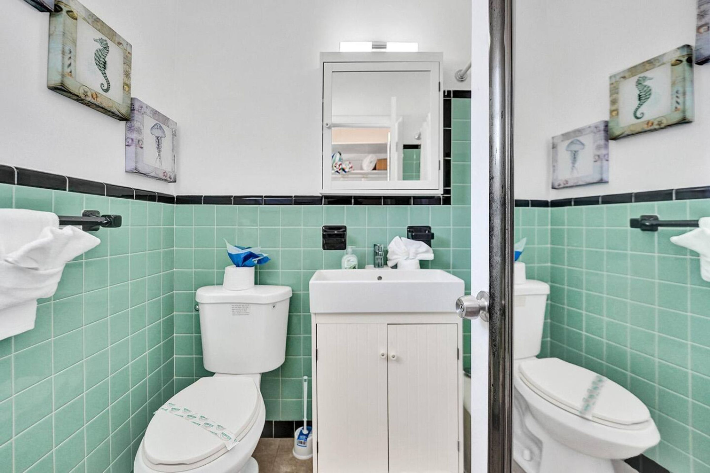 Ein Badezimmer mit grün gefliesten Wänden, zwei Toiletten, einem Waschbecken und einem Schrank.