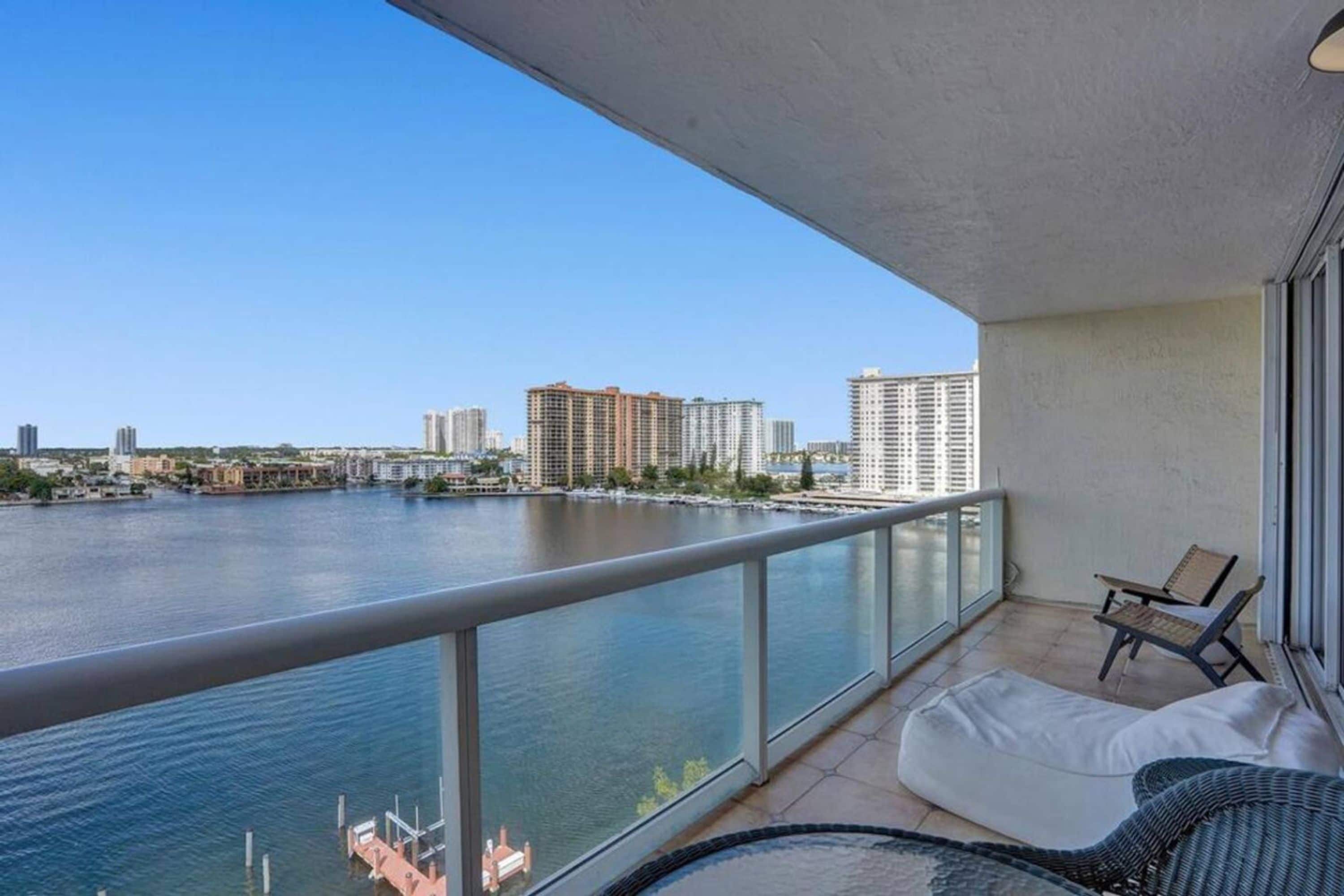 Un balcón con vista a un río y un paisaje urbano, que incluye una zona de estar con cojines y una mesa.