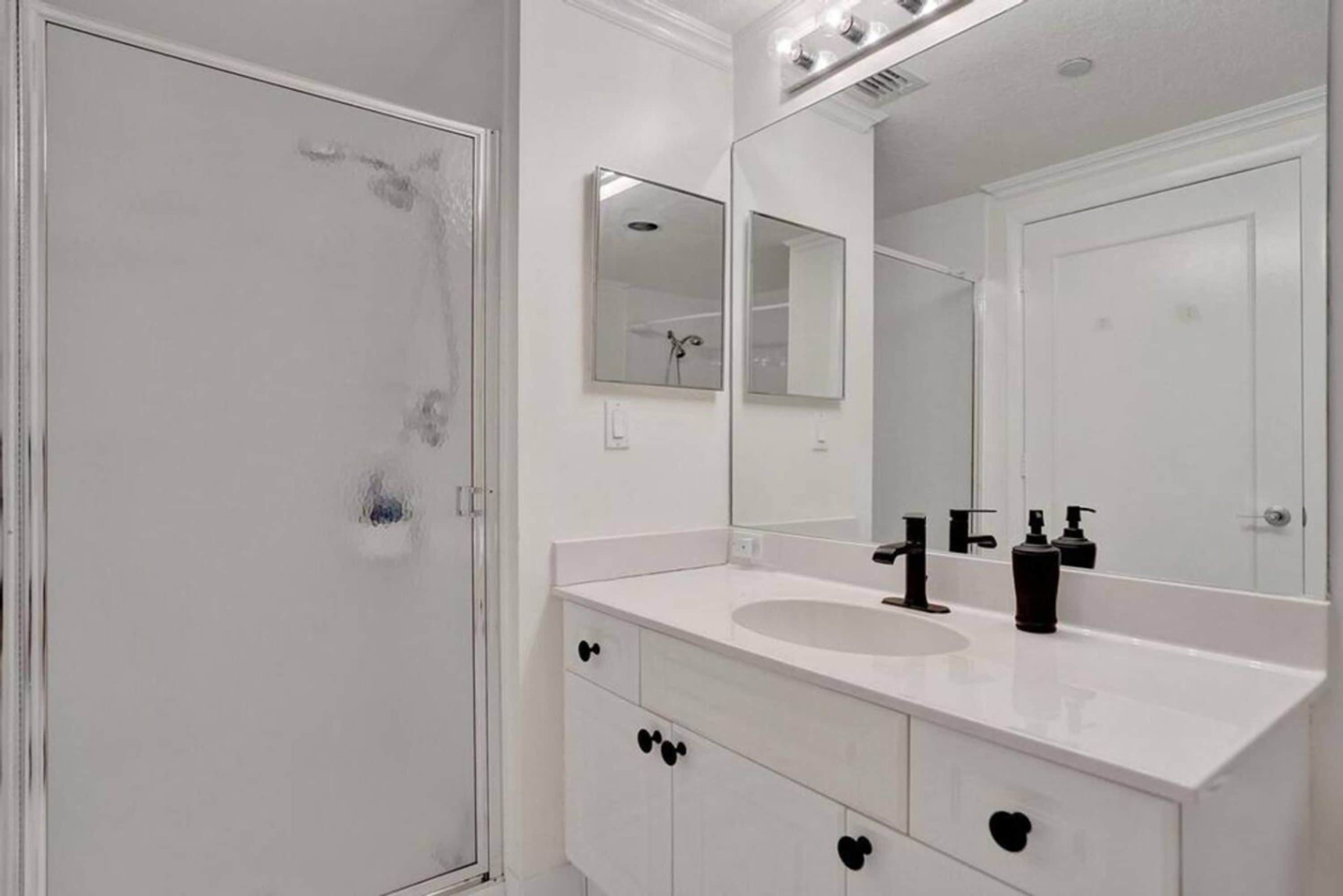 Une salle de bain avec un meuble-lavabo blanc, un évier, un miroir et une porte de douche.