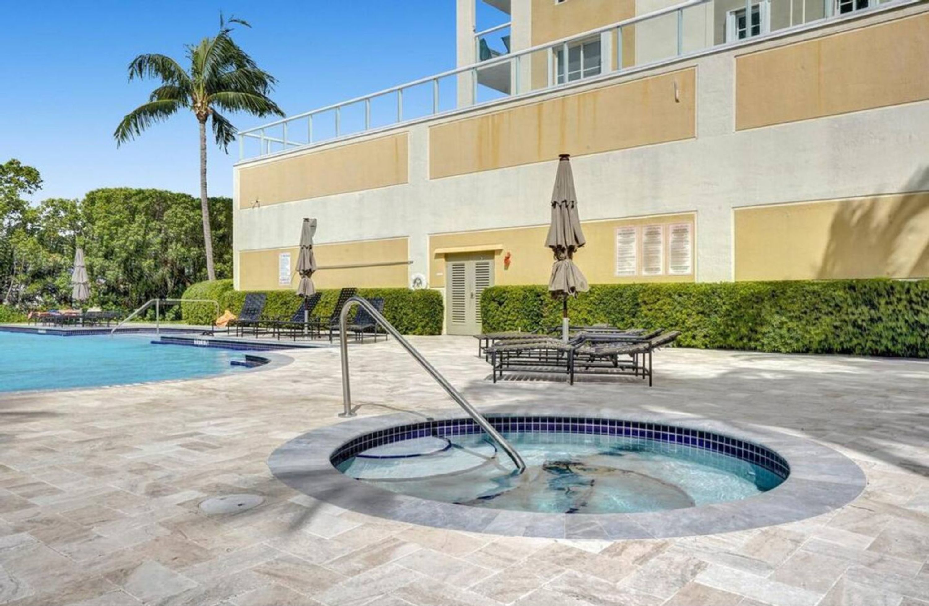 A swimming pool area with lounge chairs and umbrellas, a hot tub, and a building with a balcony.