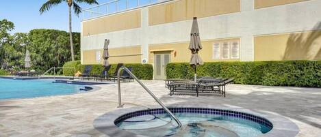 A swimming pool area with lounge chairs and umbrellas, a hot tub, and a building with a balcony.