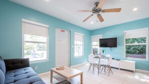 A modern living room with a sofa, a coffee table, a dining area with chairs, a TV mounted on the wall, and a ceiling fan. - Casa Del Mar Deluxe Studio 2 (Hollywood)